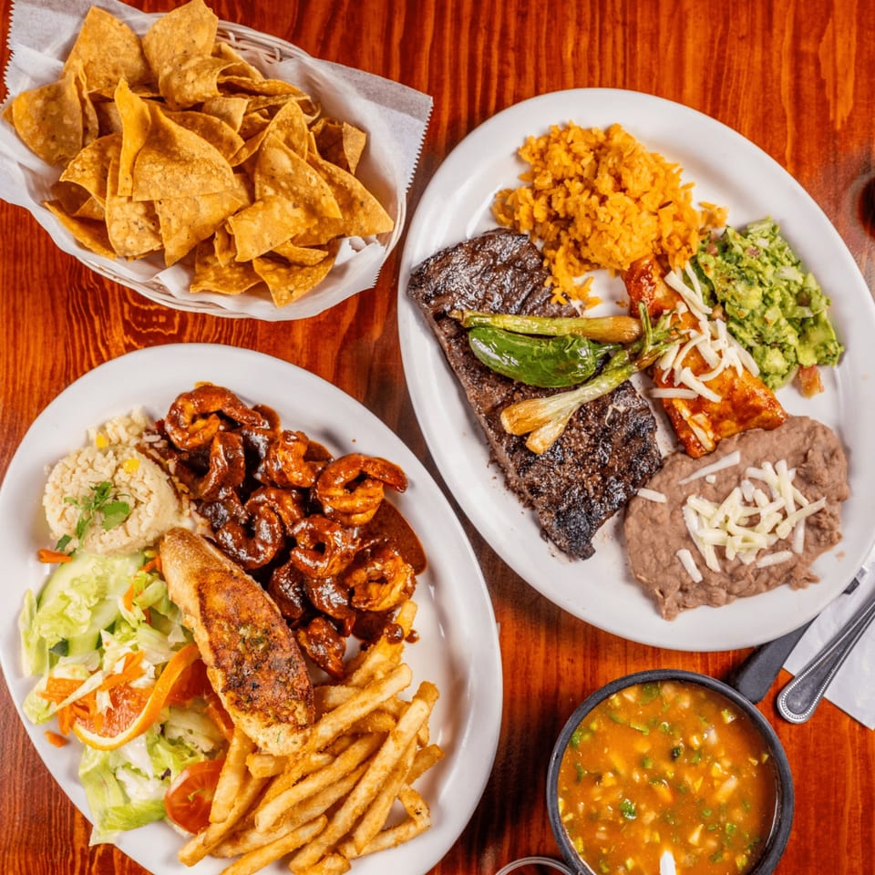 Two plates of Mexican food, including steak, shrimp, rice, beans, guacamole, and chips on a wooden table.