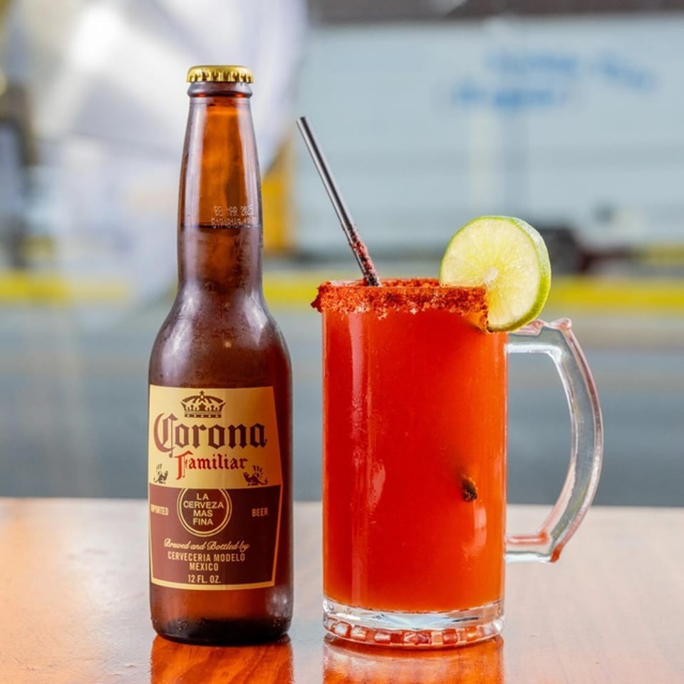 A Corona Familiar bottle next to a michelada with a lime wedge and chili-salted rim.