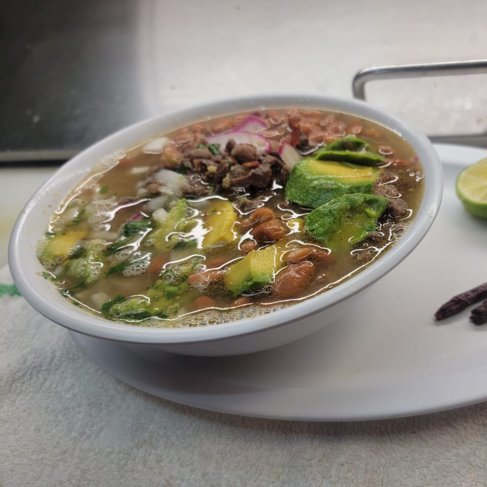 A white bowl of Mexican soup with beans, avocado, and meat, served with lime.