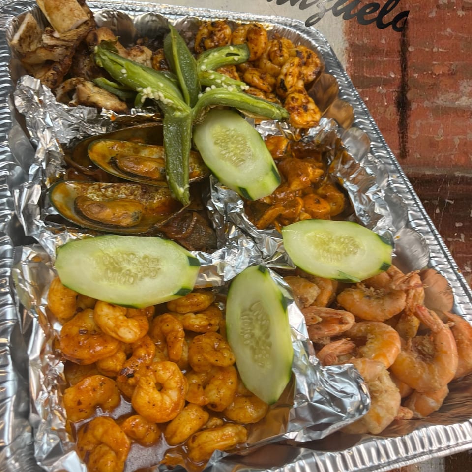 A foil tray with grilled chicken, mussels, shrimp, jalapeños, and cucumber slices from El Anzuelo.